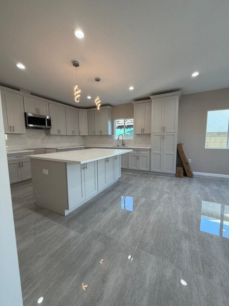 Modern kitchen with light gray cabinets, a large central island, marble countertops, and stainless steel appliances—perfect for ADU Homes in San Pedro. Large windows and pendant lights add to the bright, spacious feel.