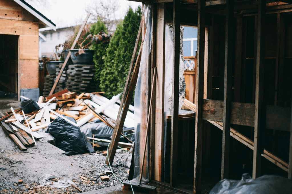 A partially constructed or demolished wooden structure with exposed framing is visible in the foreground. Nearby, black garbage bags and construction debris are scattered on gravel, reflecting ongoing work to meet California ADU laws amidst 2026 updates.