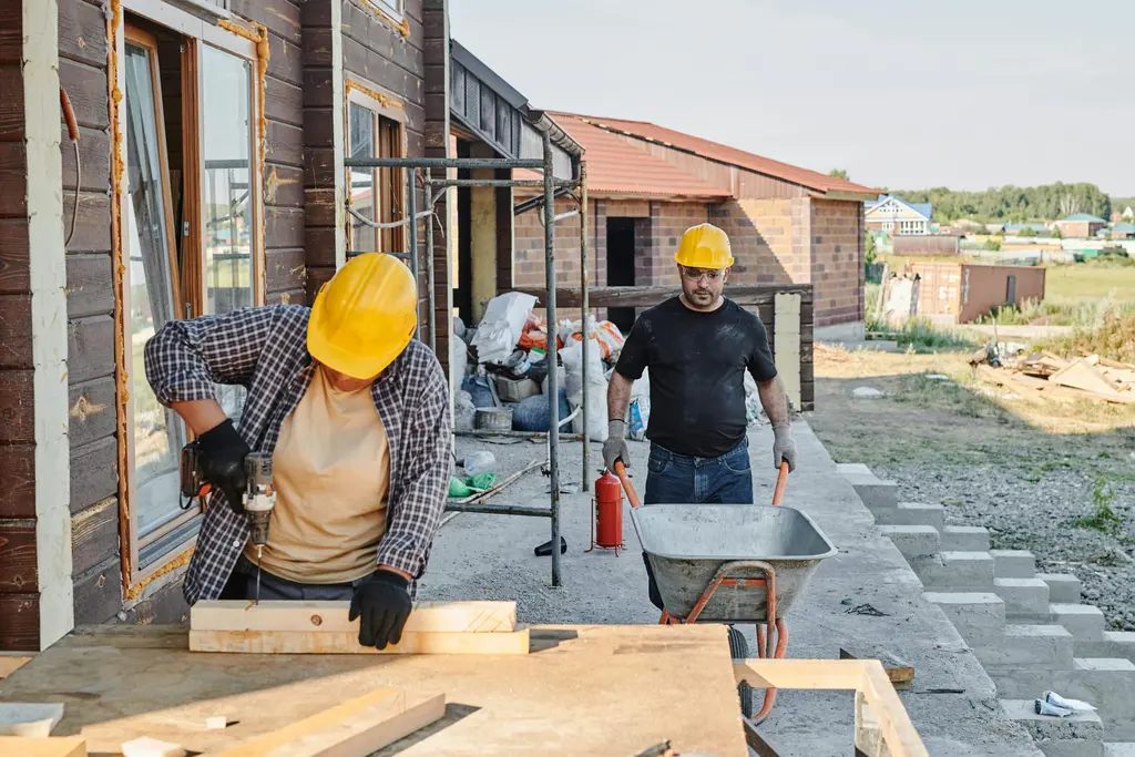 Two construction workers wearing yellow hard hats work outside a building. One uses a drill on a wooden plank at a table, while the other stands behind holding a wheelbarrow. Building materials and tools are scattered around the outdoor worksite.