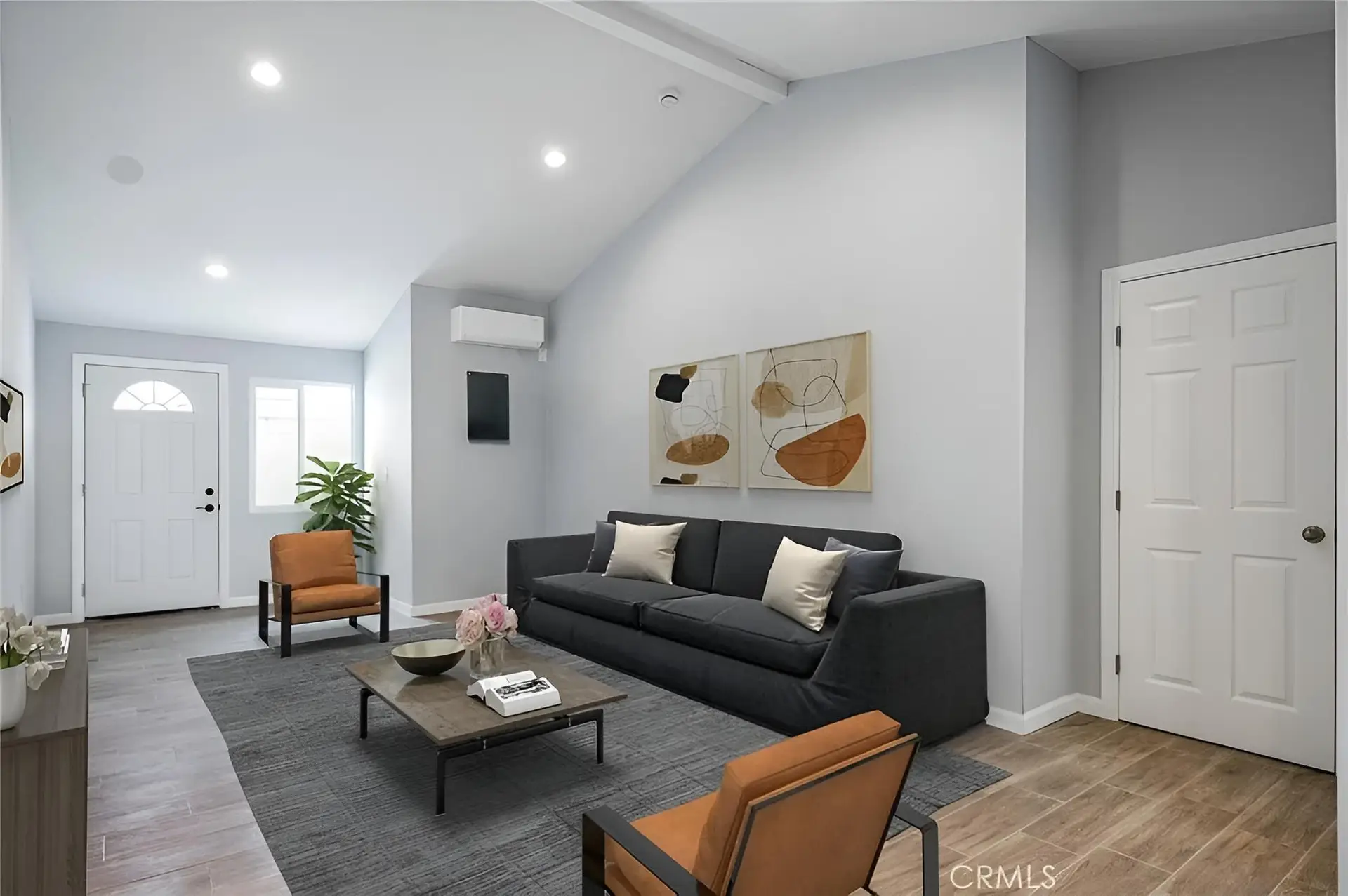 Modern living room with gray walls, a black sofa with beige pillows, two orange armchairs, a gray rug, and a wooden coffee table with books and flowers. Two abstract art pieces hang above the sofa. Light wood floor and a plant by the door add warmth—an inspiring space that showcases the thoughtful design often seen in ADU San Pedro interiors or homes completed after kitchen and bathroom remodeling.