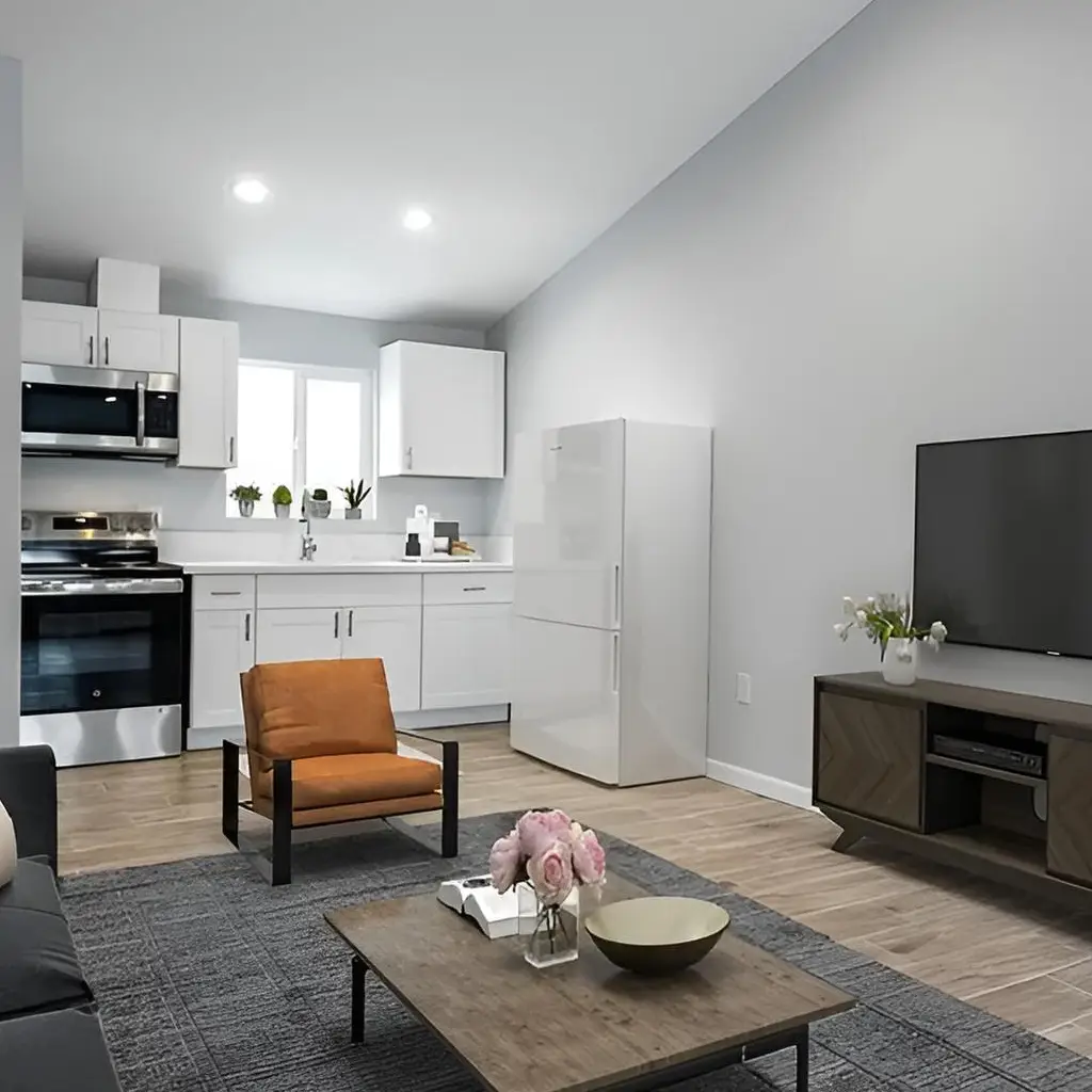Modern, bright studio ADU San Pedro with a kitchen, TV, orange armchair, gray sofa, and coffee table with books and flowers. White cabinets, stainless stove, fridge, plants, hardwood floor, and gray rug emphasize a clean, minimalist style—perfect for those seeking new construction or expert kitchen and bathroom remodeling.